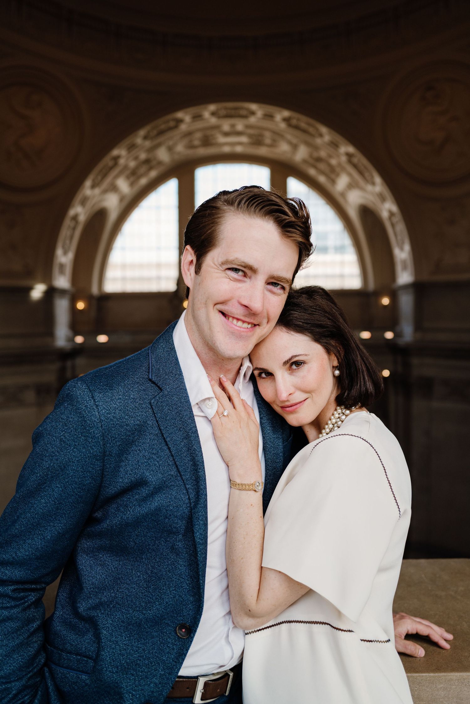 A couple in classic wedding attire shares intimate moments in an elegant indoor setting with arched architectural details.