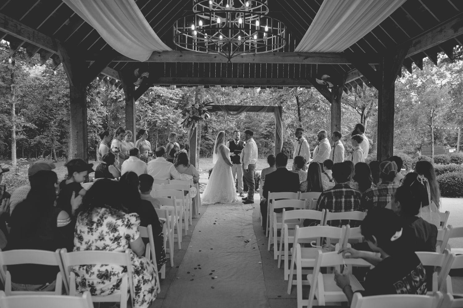 A couple in romantic wedding attire shares tender moments in a rustic outdoor setting with wood buildings and green trees.