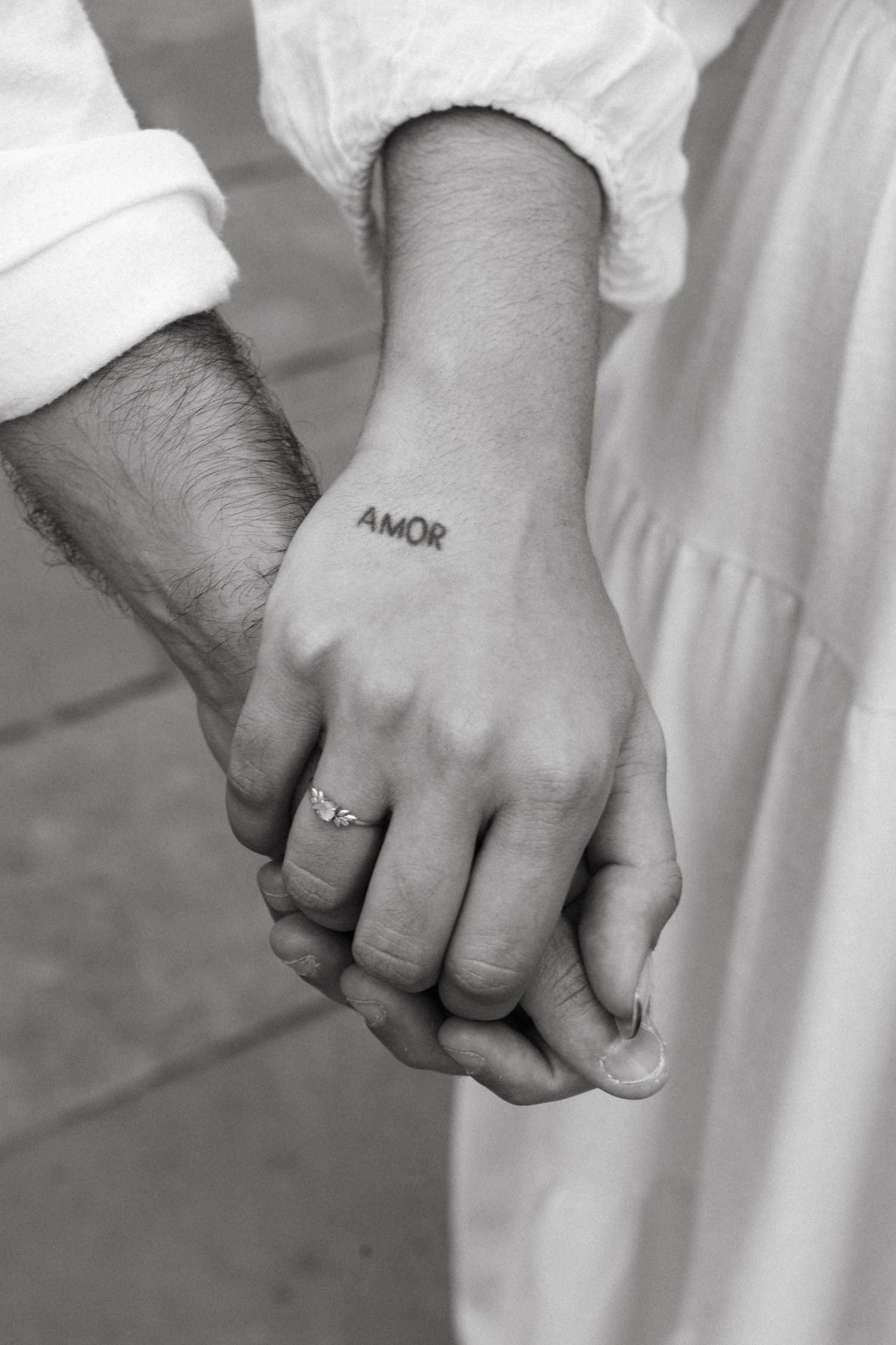 Close-up of joined hands showing a tattoo reading 'amor' and wedding rings.