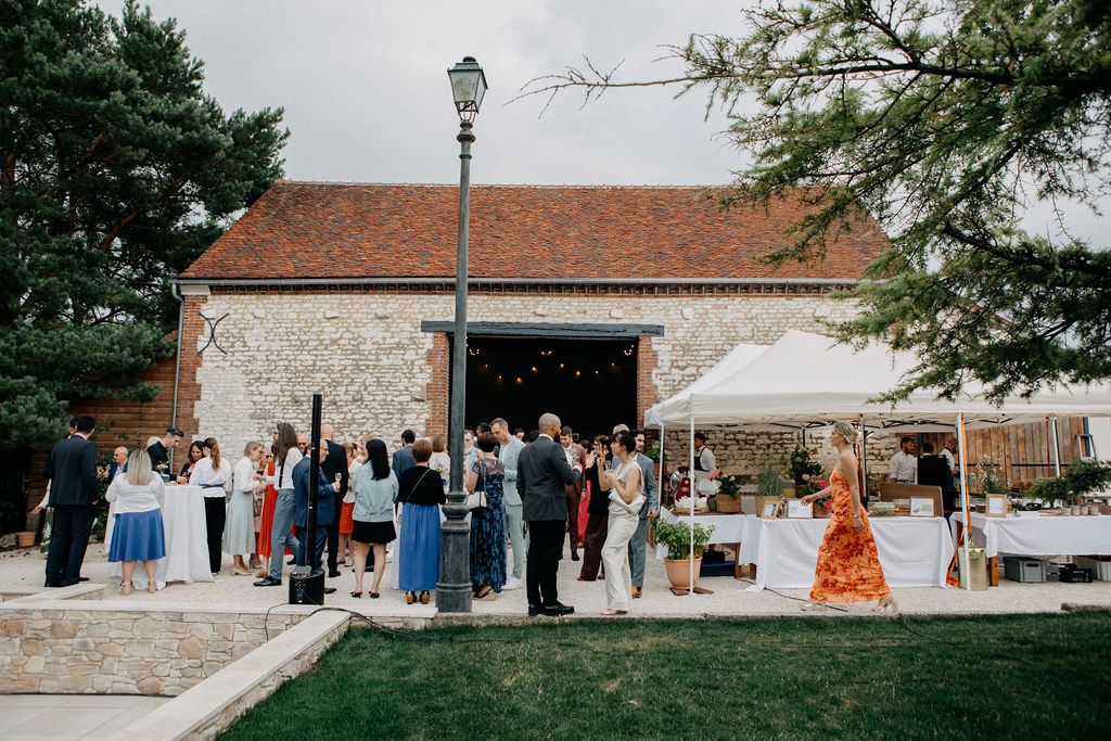 Elegantly dressed wedding guests mingle outside a charming rustic barn venue on a sunny day.