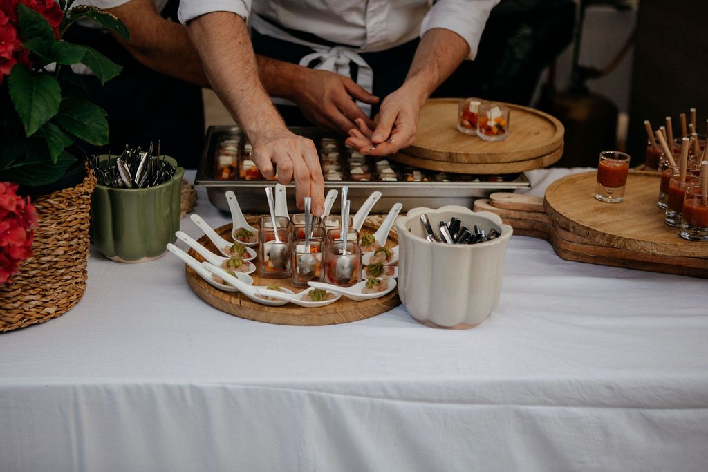 A caterer arranges gourmet wedding appetizers on wooden boards at a white-clothed catering table.