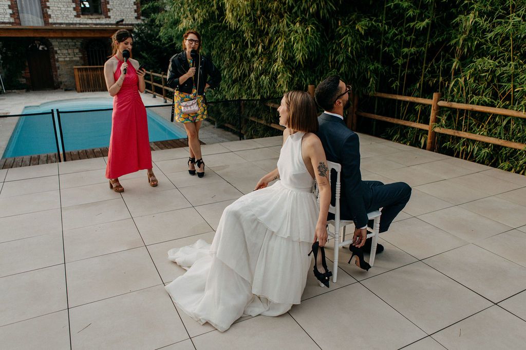 A bride in a white gown sits with her groom while a guest in red speaks at their wedding.