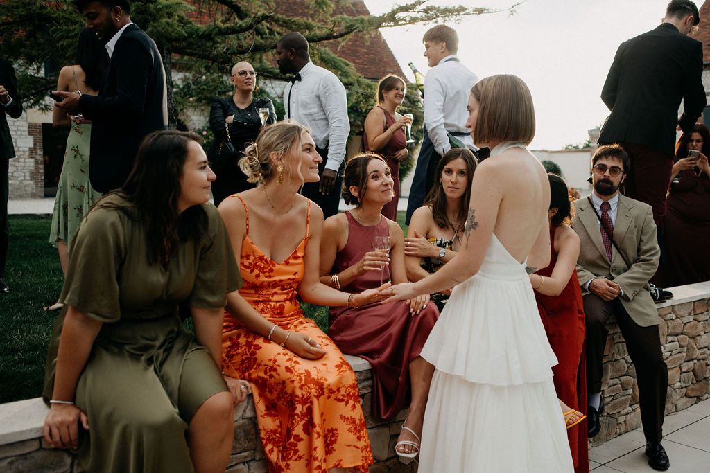 A bride in a white dress chats with female wedding guests in colorful dresses at an outdoor reception.