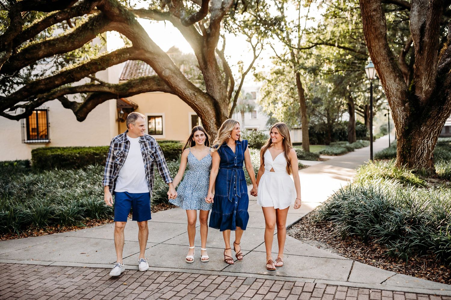 A group in casual summer attire walks together on a sunlit brick pathway under large trees near a building.