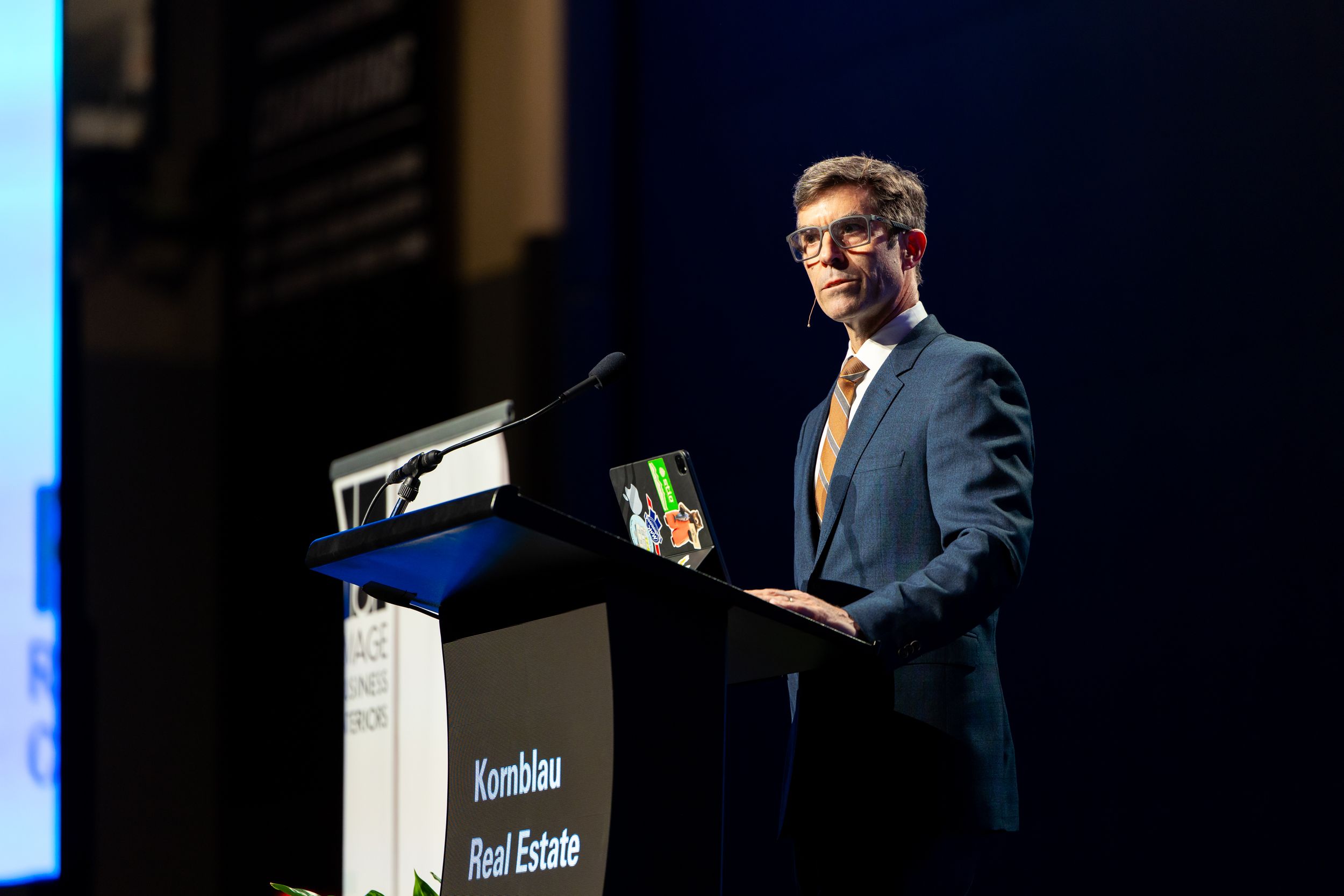 Man in a navy suit and glasses speaks at a podium with a Kombau Real Estate sign during a conference presentation.