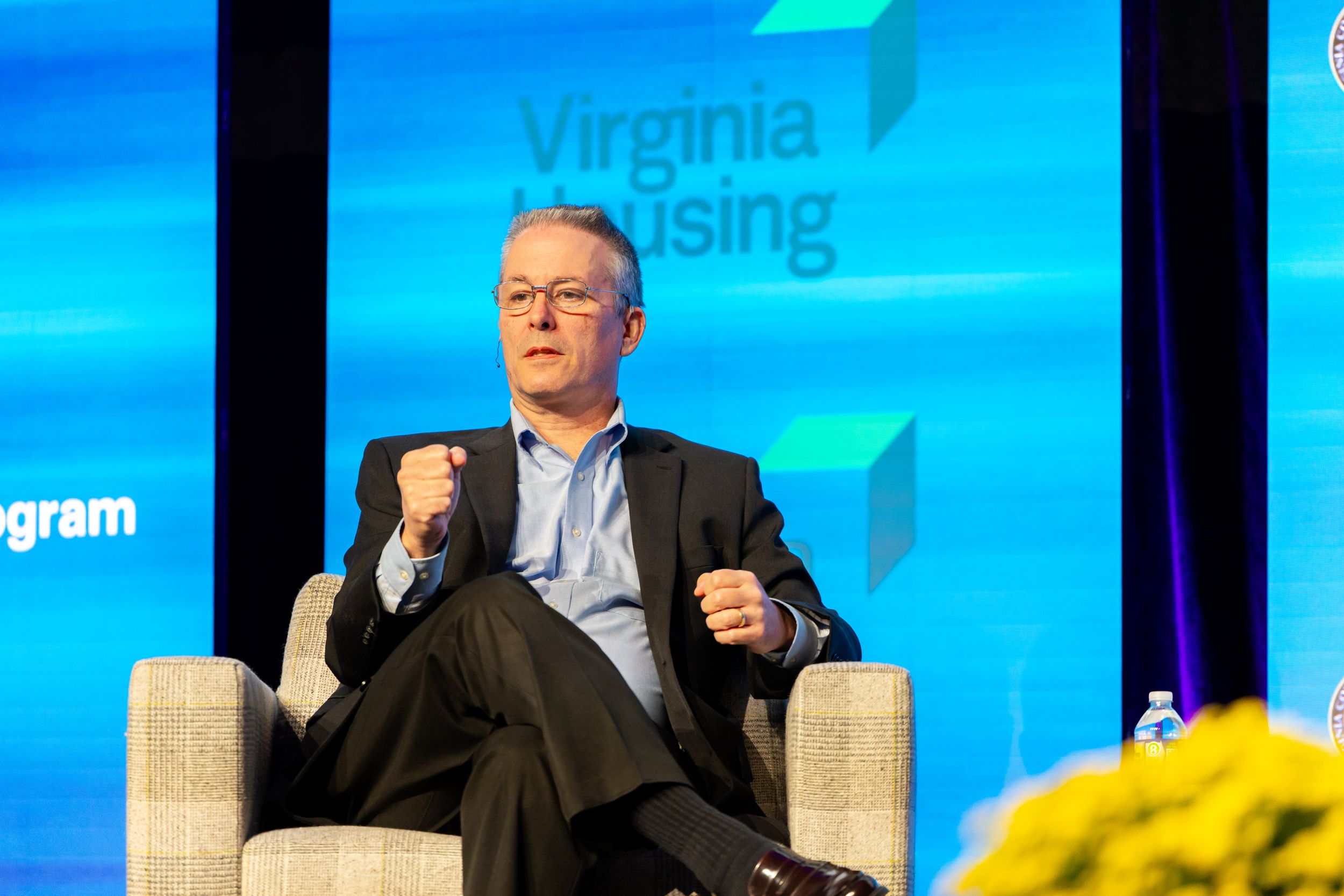 A man in glasses and a dark blazer gestures while speaking from an armchair on stage at a Virginia Housing event.