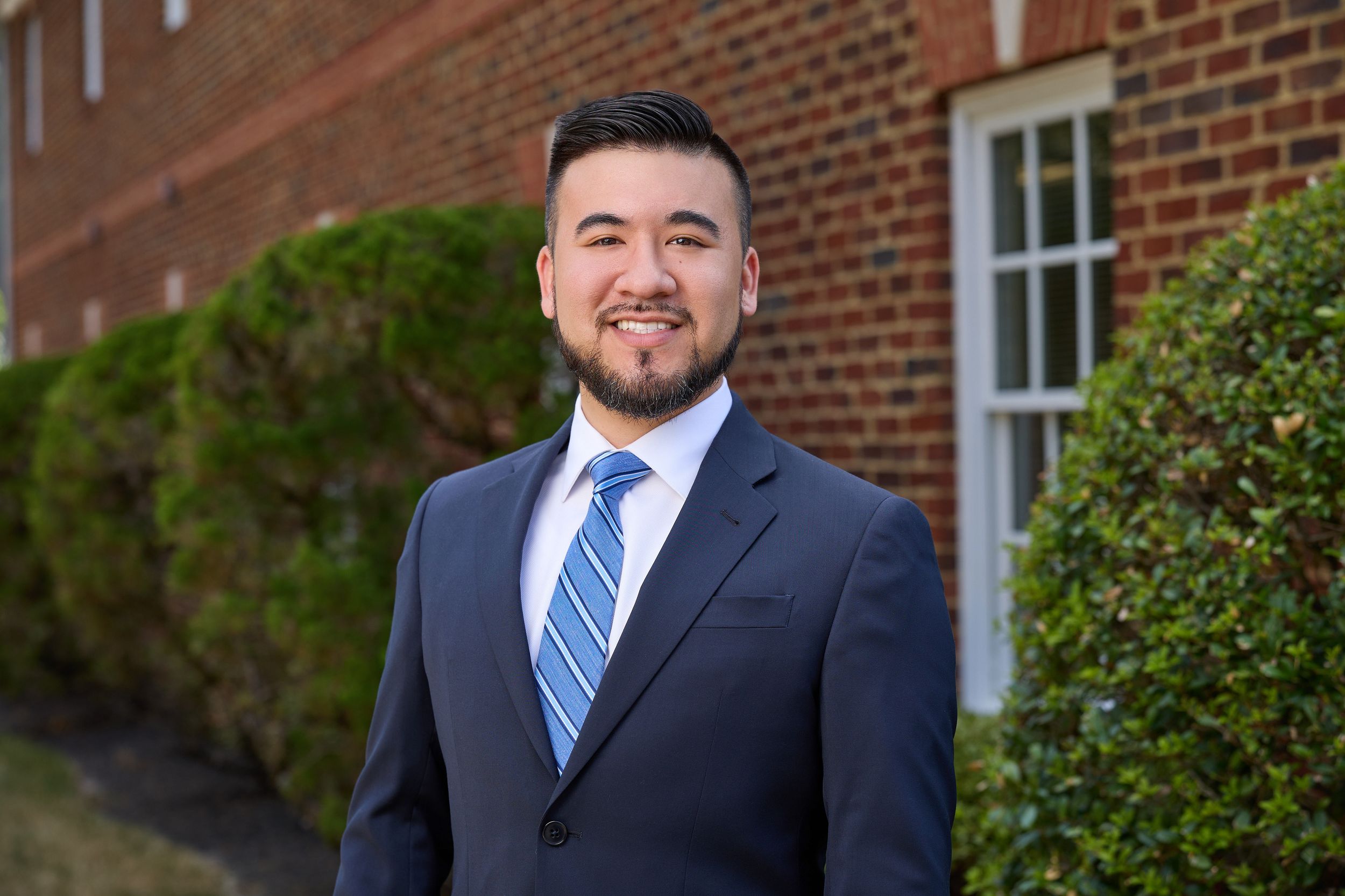 Professional headshot portrait against a brick building exterior with windows and greenery in the background.