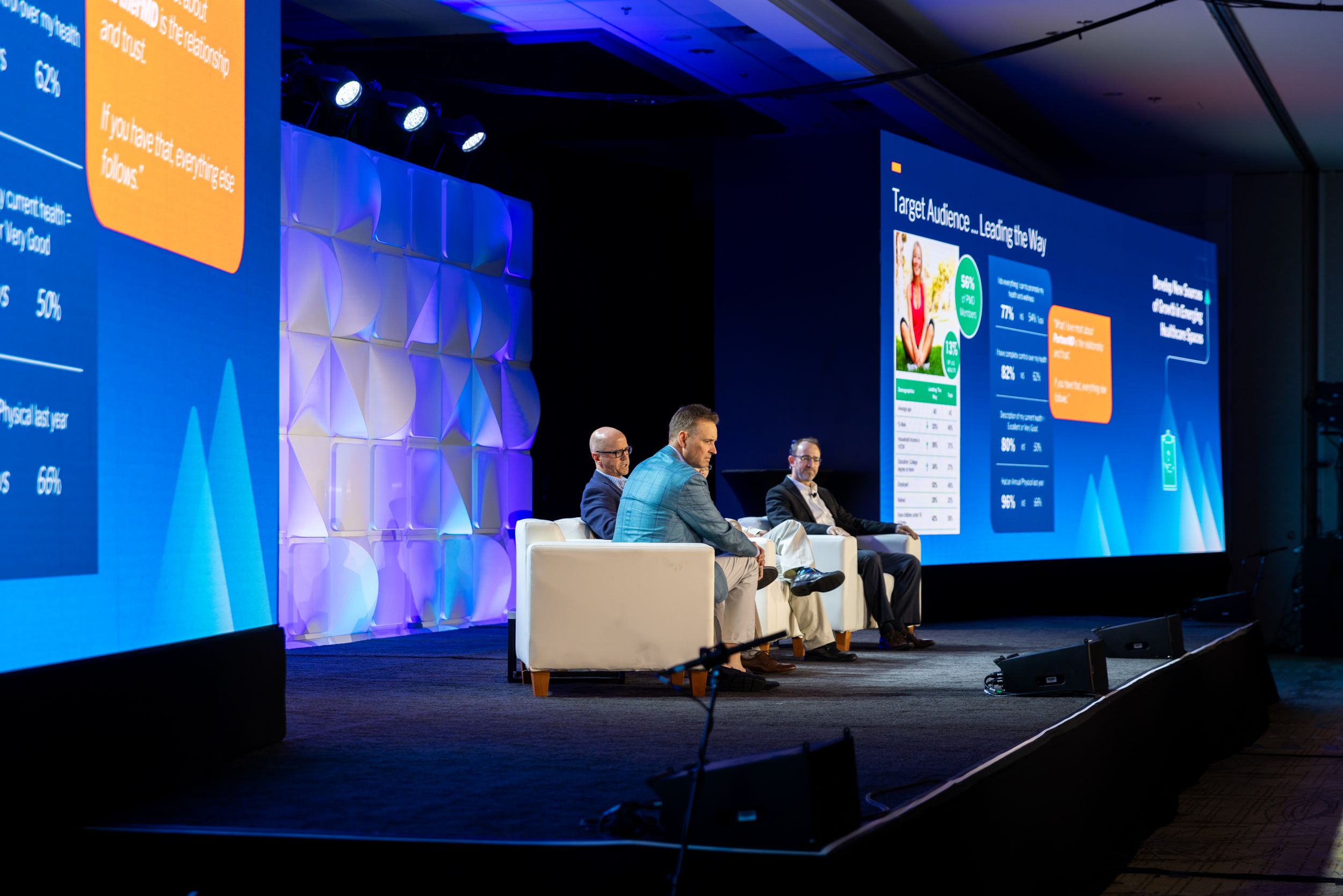 Three speakers sit on white armchairs on a conference stage, flanked by large illuminated screens displaying presentation slides.