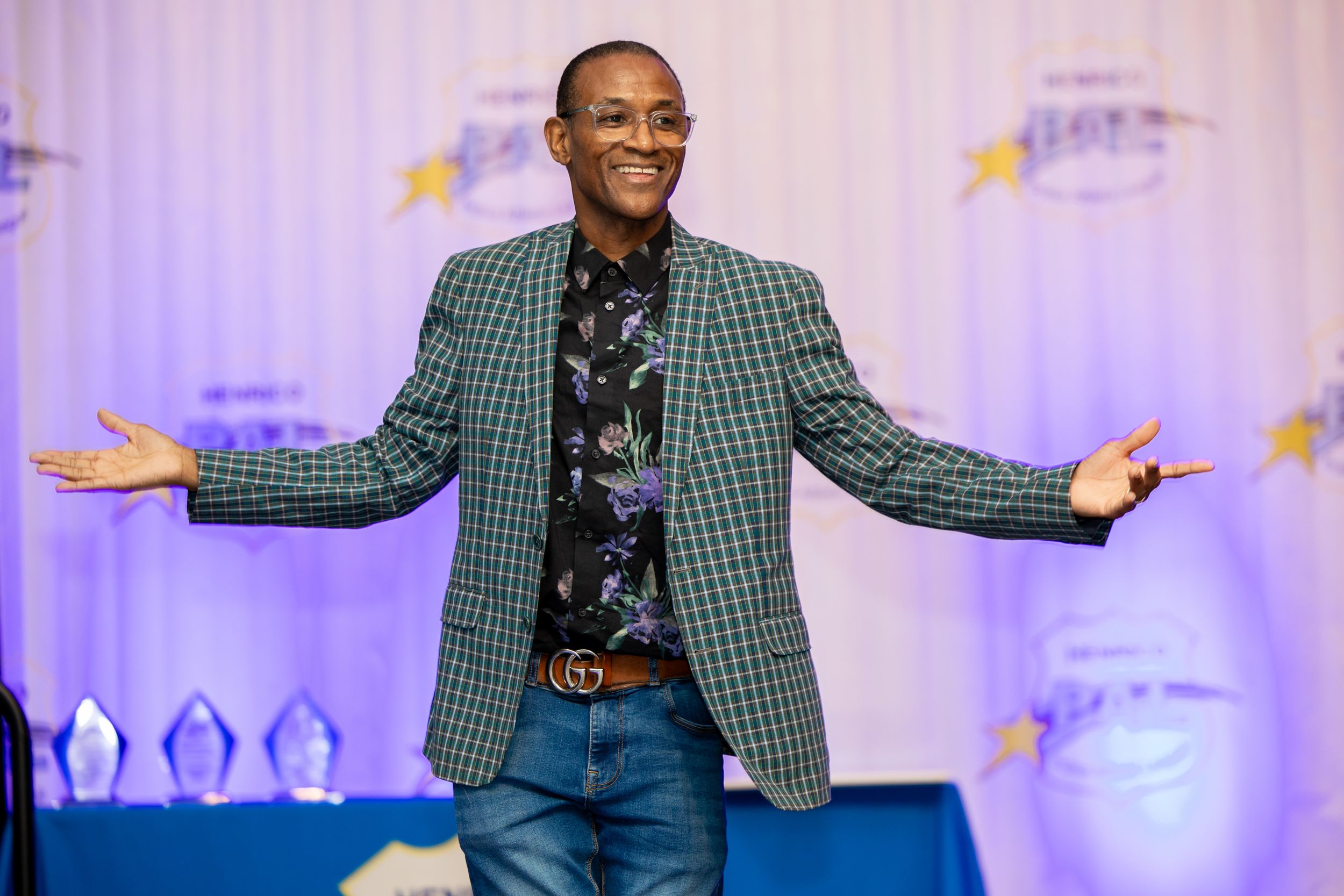 Speaker in green plaid blazer gestures energetically on stage with colorful lighting effects in the background.