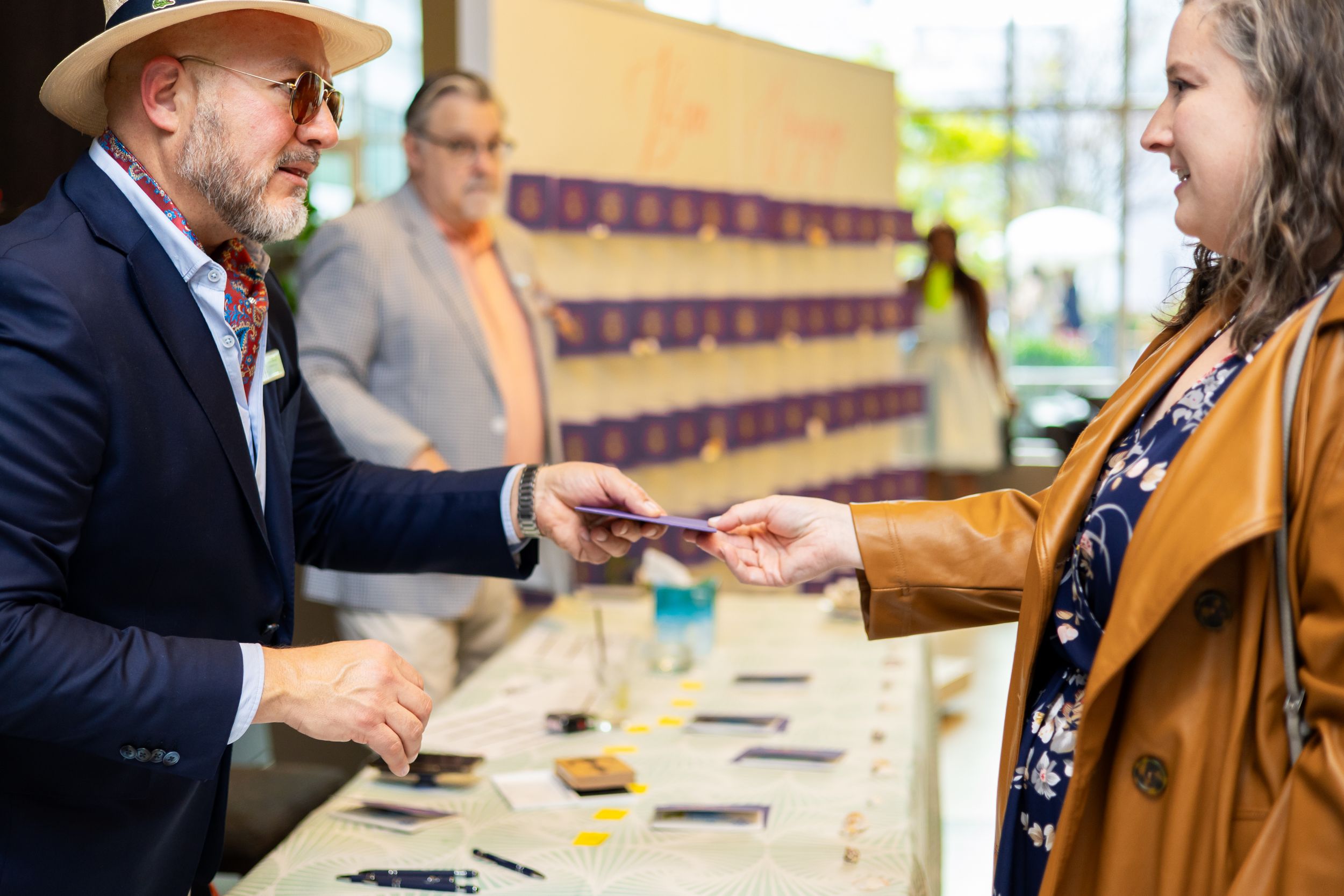 Bearded man in a straw hat and navy blazer hands a card to a smiling woman in a tan jacket at a busy event registration table.