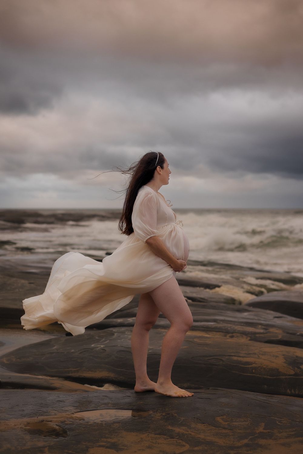 Pregnant woman standing on windswept Sunshine Coast beach during a moody maternity photoshoot with dramatic skies.