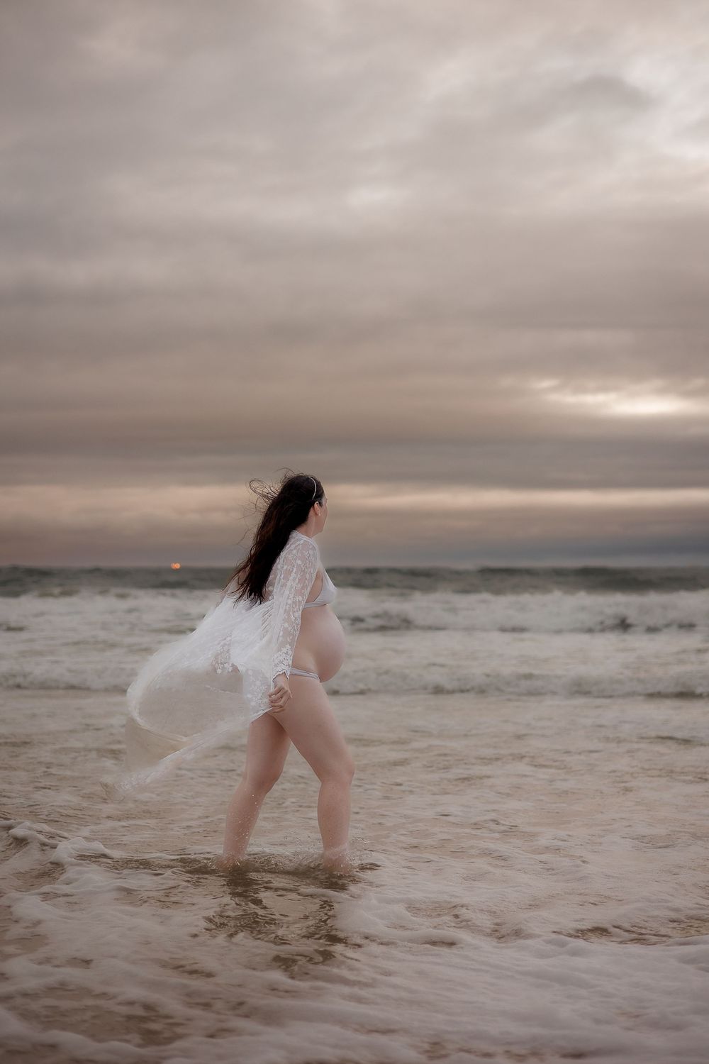 Moody maternity photoshoot at Sunshine Coast beach, pregnant woman in flowing dress under overcast skies with dramatic coastal scenery.