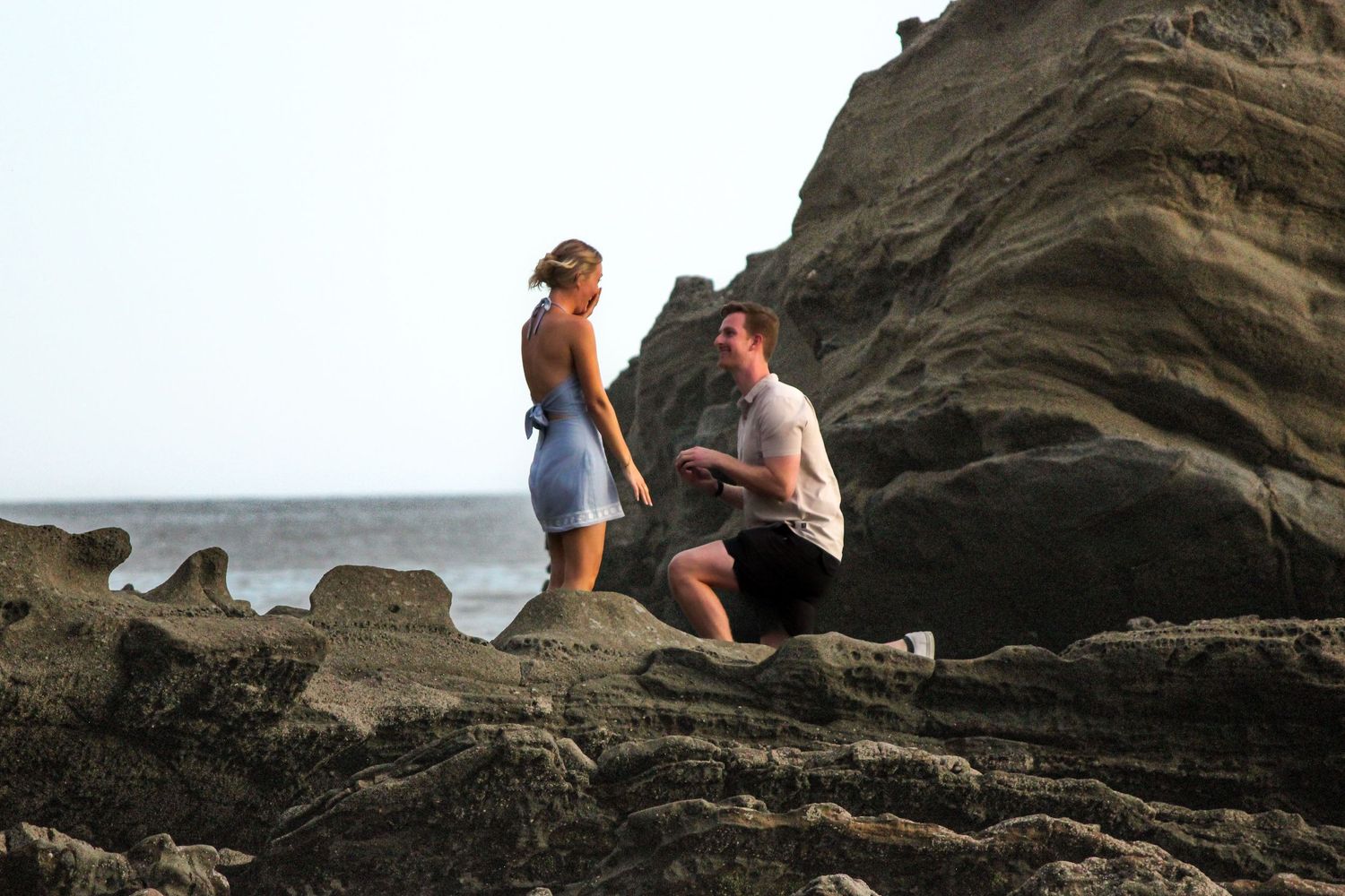 A beach engagement photoshoot taking place on rocks. Man on a knee proposing to a female in a blue dress