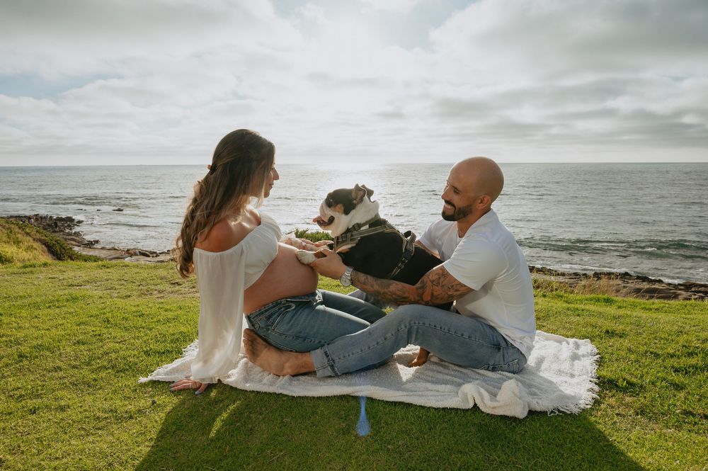 Pregnant couple sitting on a blanket by the ocean, smiling at each other with their dog between them.