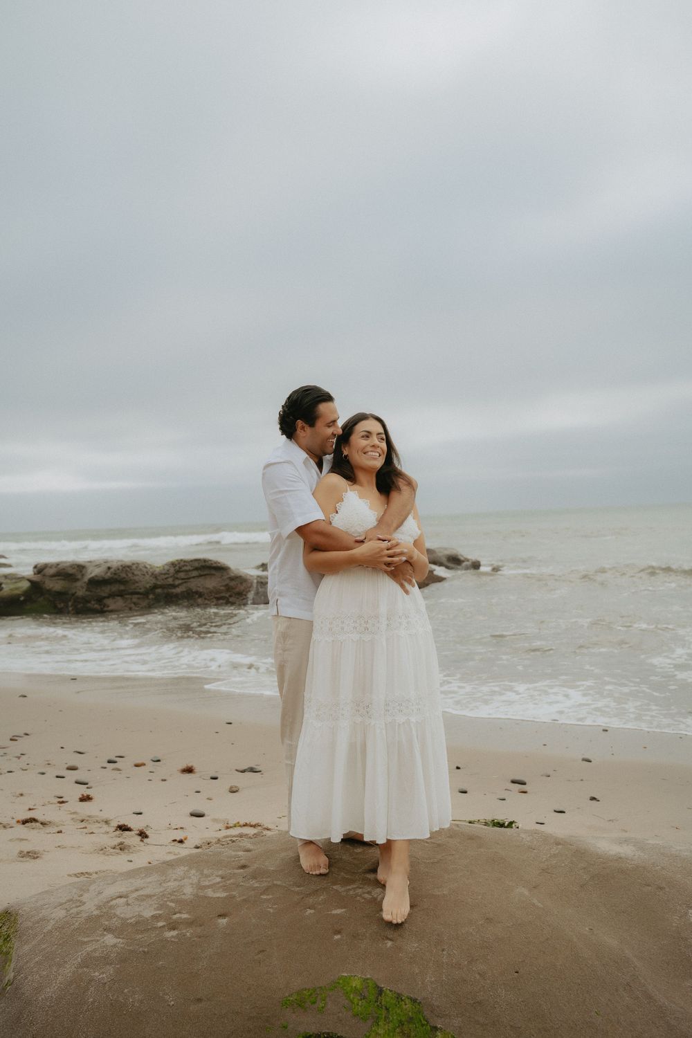 Couple embracing on a beach, with the woman in a white dress and the man standing behind her under a cloudy sky.