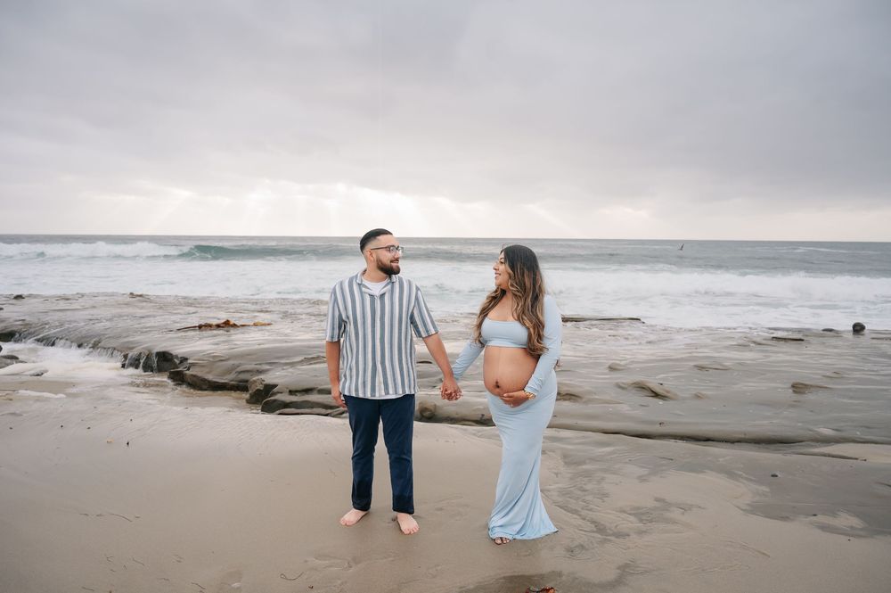 Pregnant couple holding hands on a beach, standing barefoot near the shoreline under a cloudy sky.