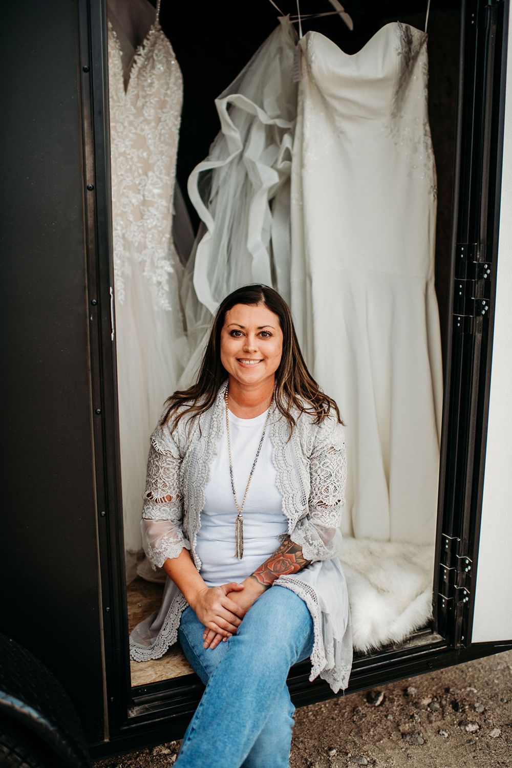 Wedding dress designer sits in front of a display of elegant white bridal gowns in a boutique.