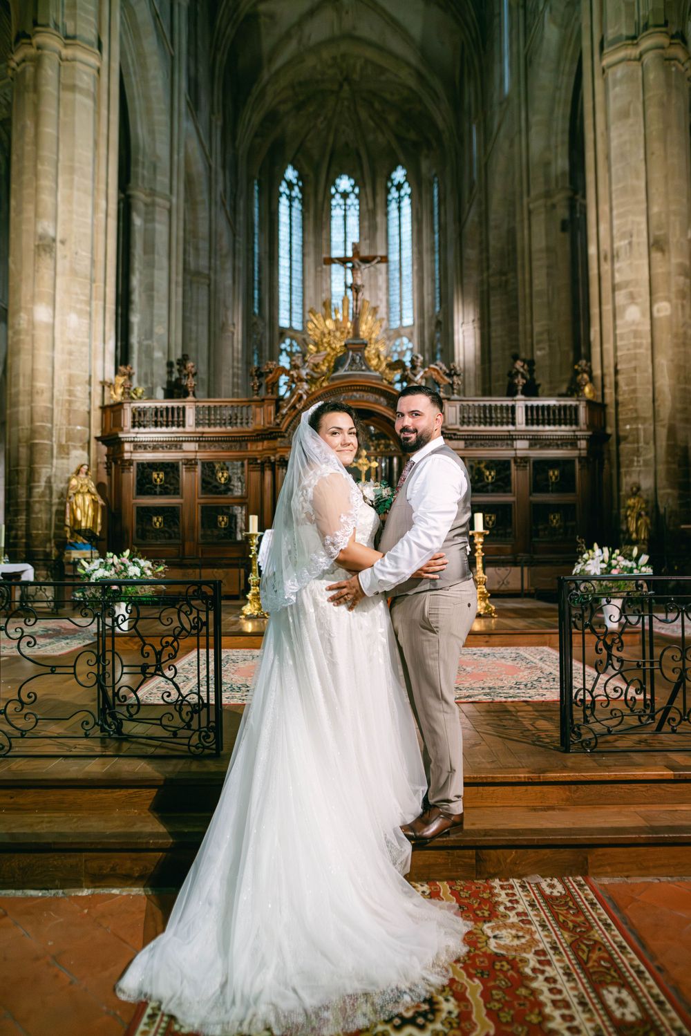 A couple poses in a grand Gothic cathedral with soaring vaulted ceilings, stained glass windows and ornate altar decorations.