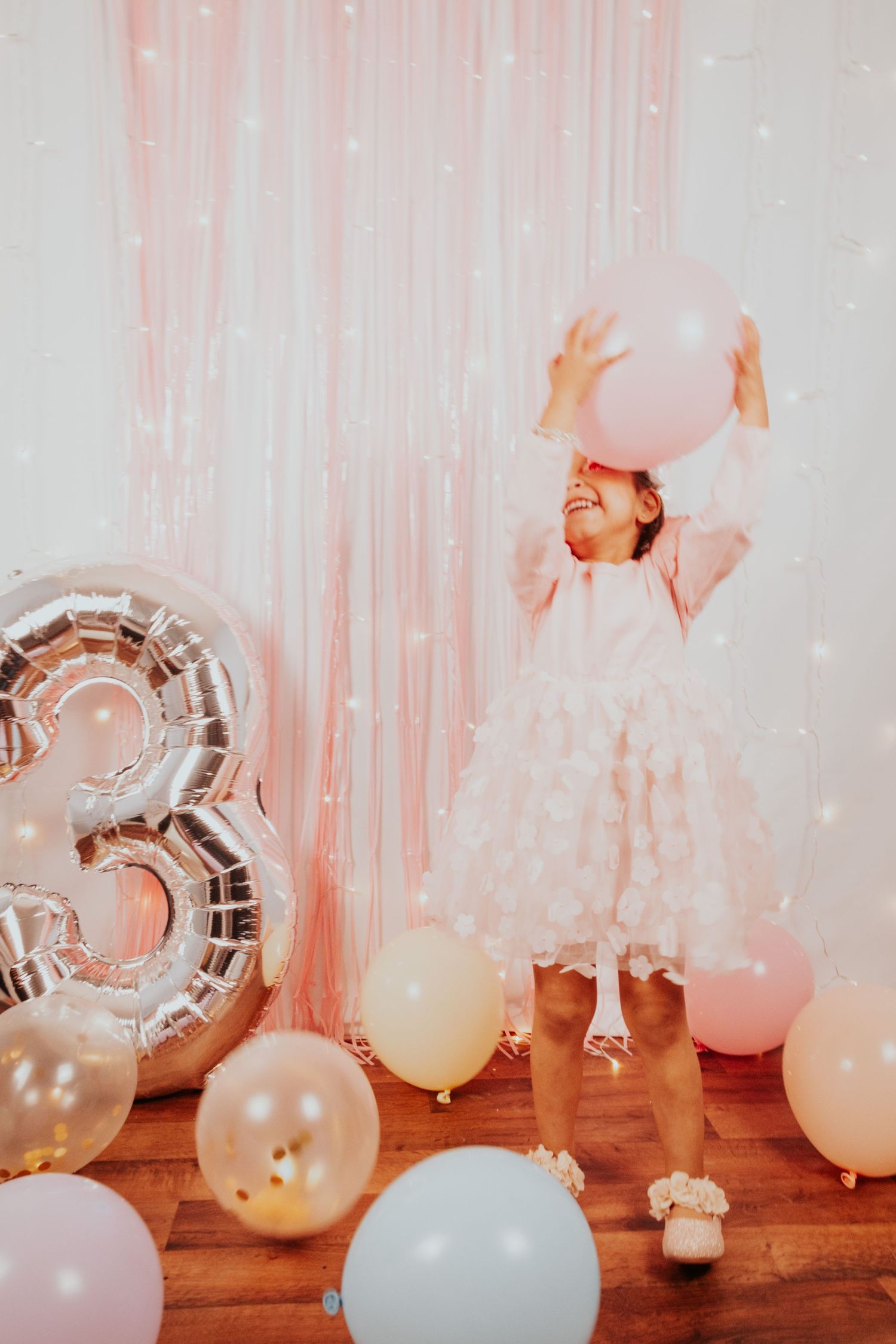 A baby wearing white and a tiny crown sits on hardwood flooring during a photoshoot.