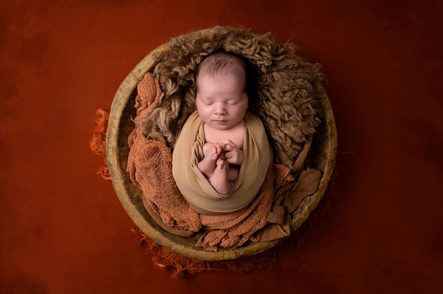 Sleeping newborn wrapped in brown fabric nestled in a rustic wooden bowl against dark background.