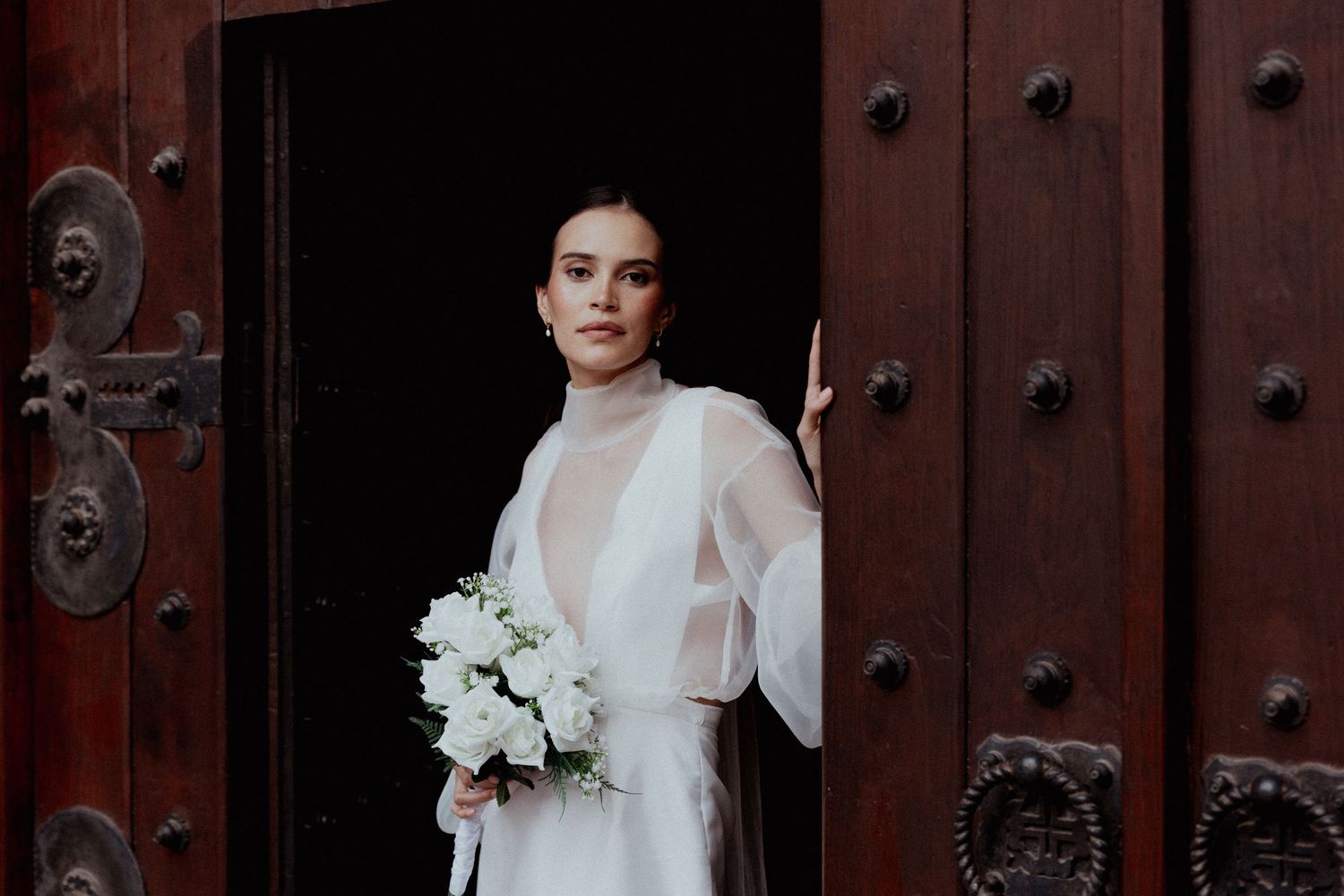 Beautiful bride in flowing white wedding dress poses elegantly against ornate dark wooden doors.