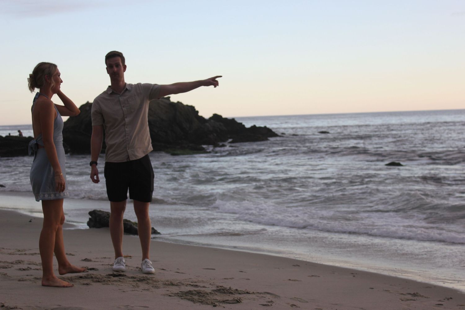 During a photoshoot a couple stands on the shore of a beach while the man points out to the horizon. There is a rocky shoreline at sunset.