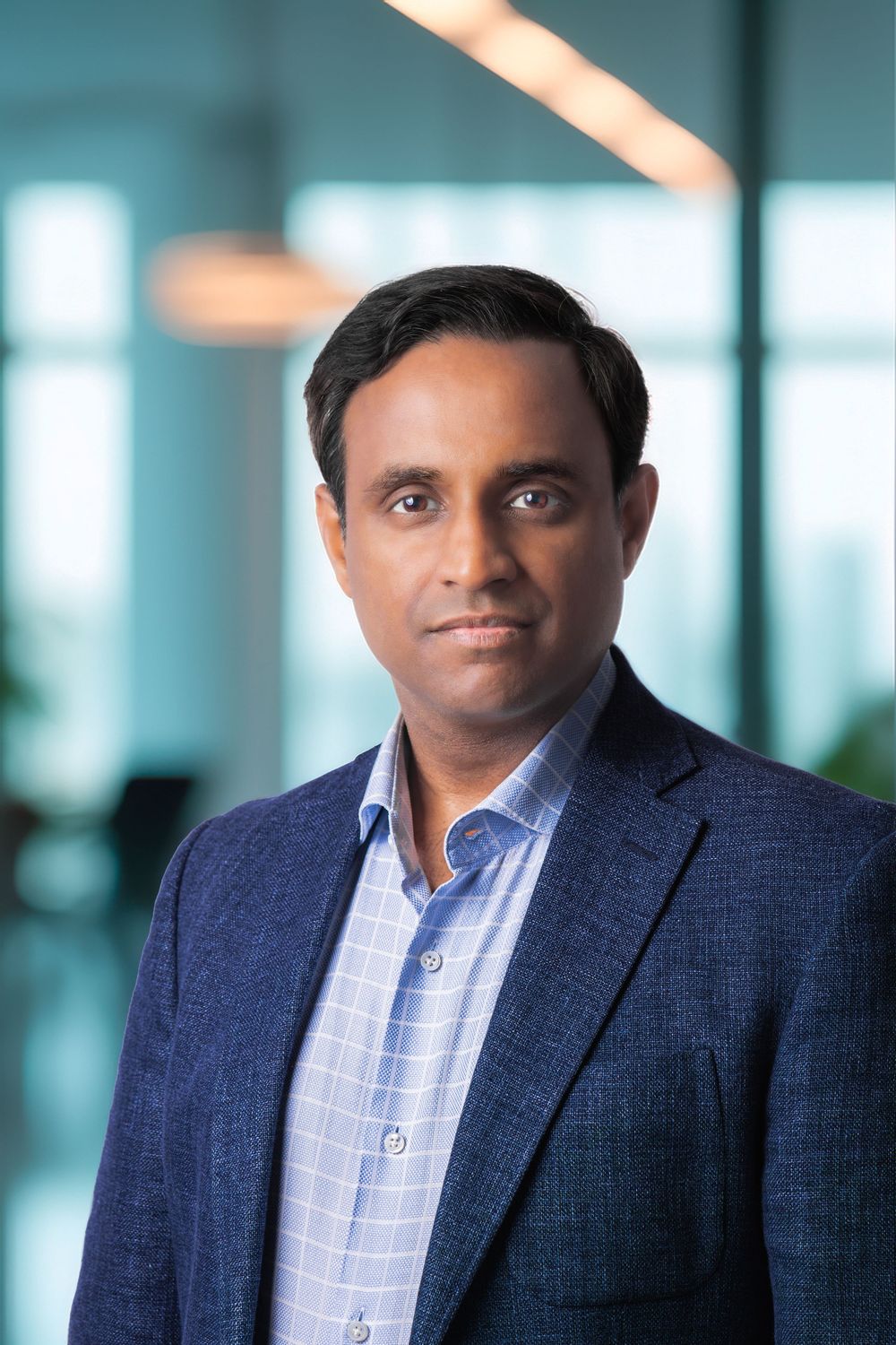 Professional headshot of a man in a blue blazer and checkered shirt in a modern office setting.