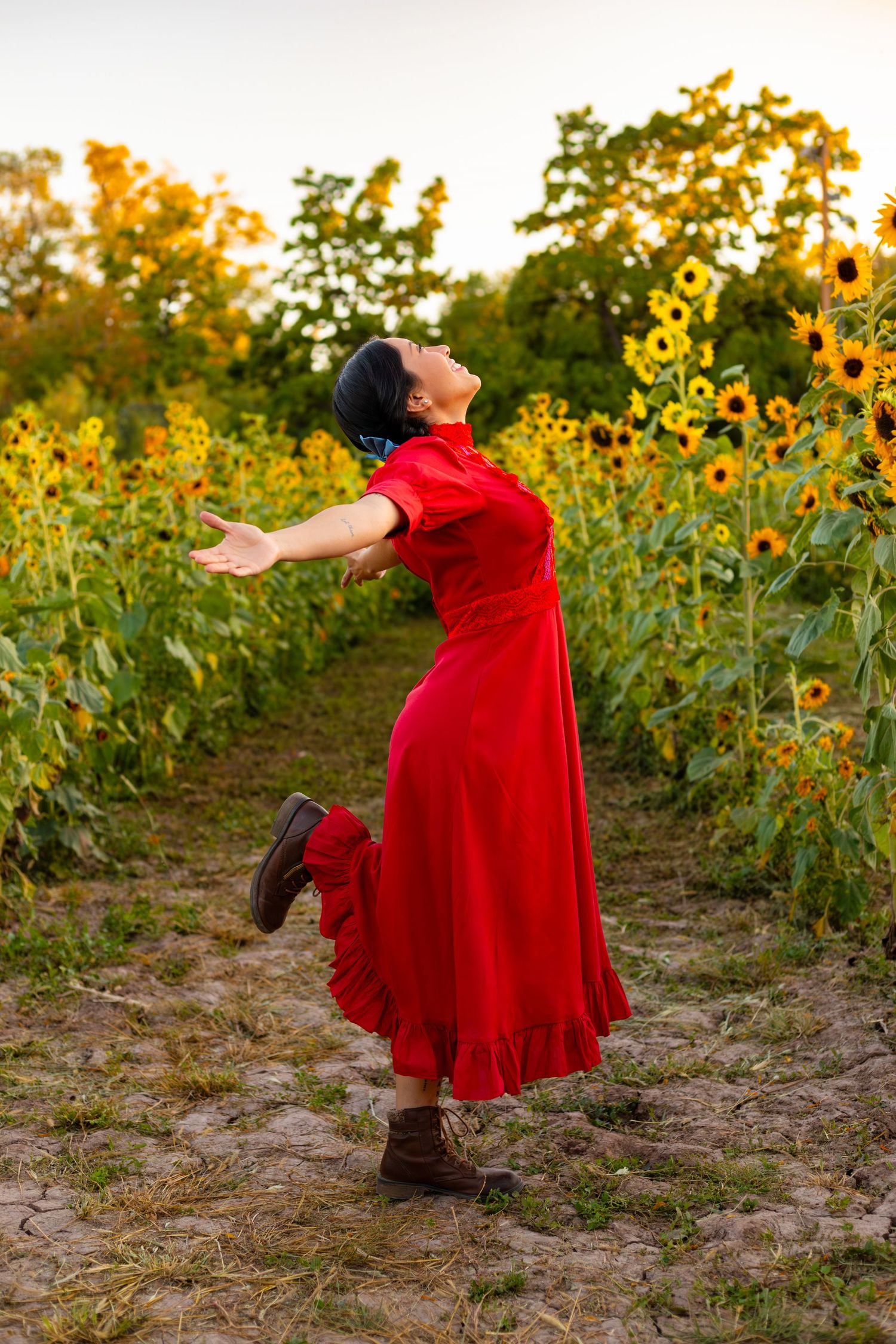 Woman in red dress joyfully poses among tall sunflowers at sunset.