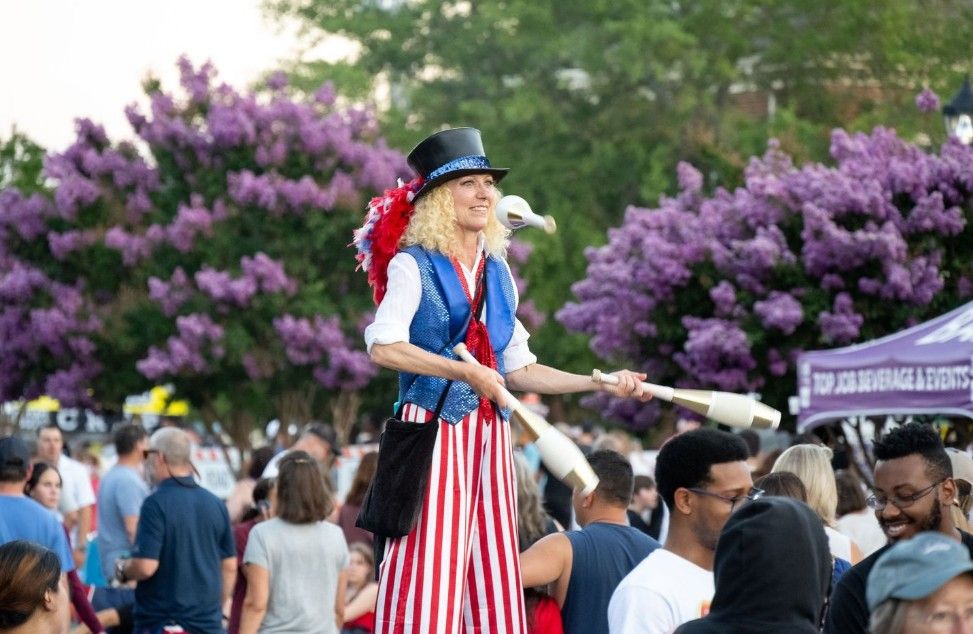 A performer in patriotic American flag attire entertaining a crowd in front of blooming purple lilac bushes.