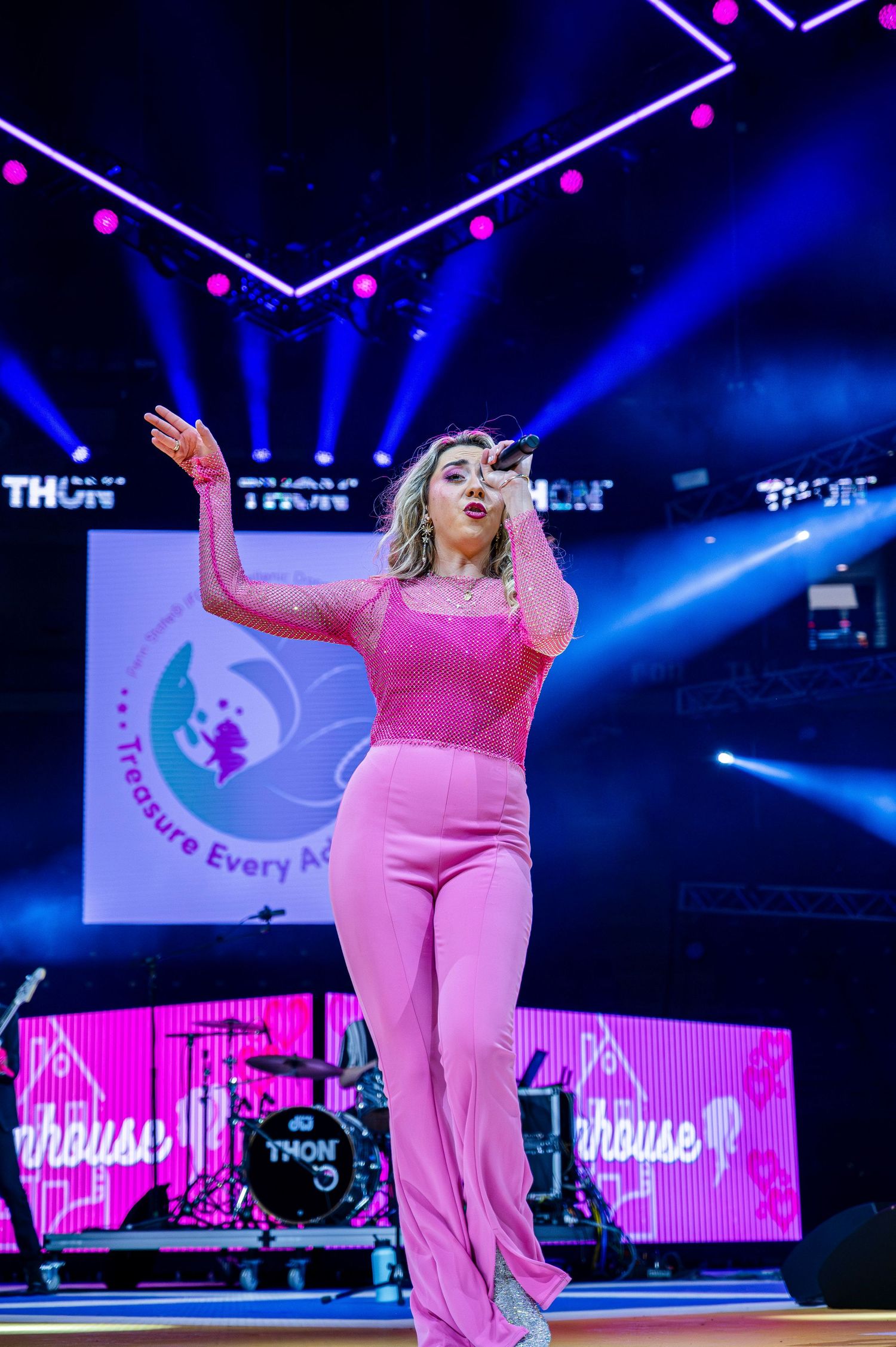 A performer in pink attire energetically sings on stage with dramatic blue lighting and digital displays in the background.