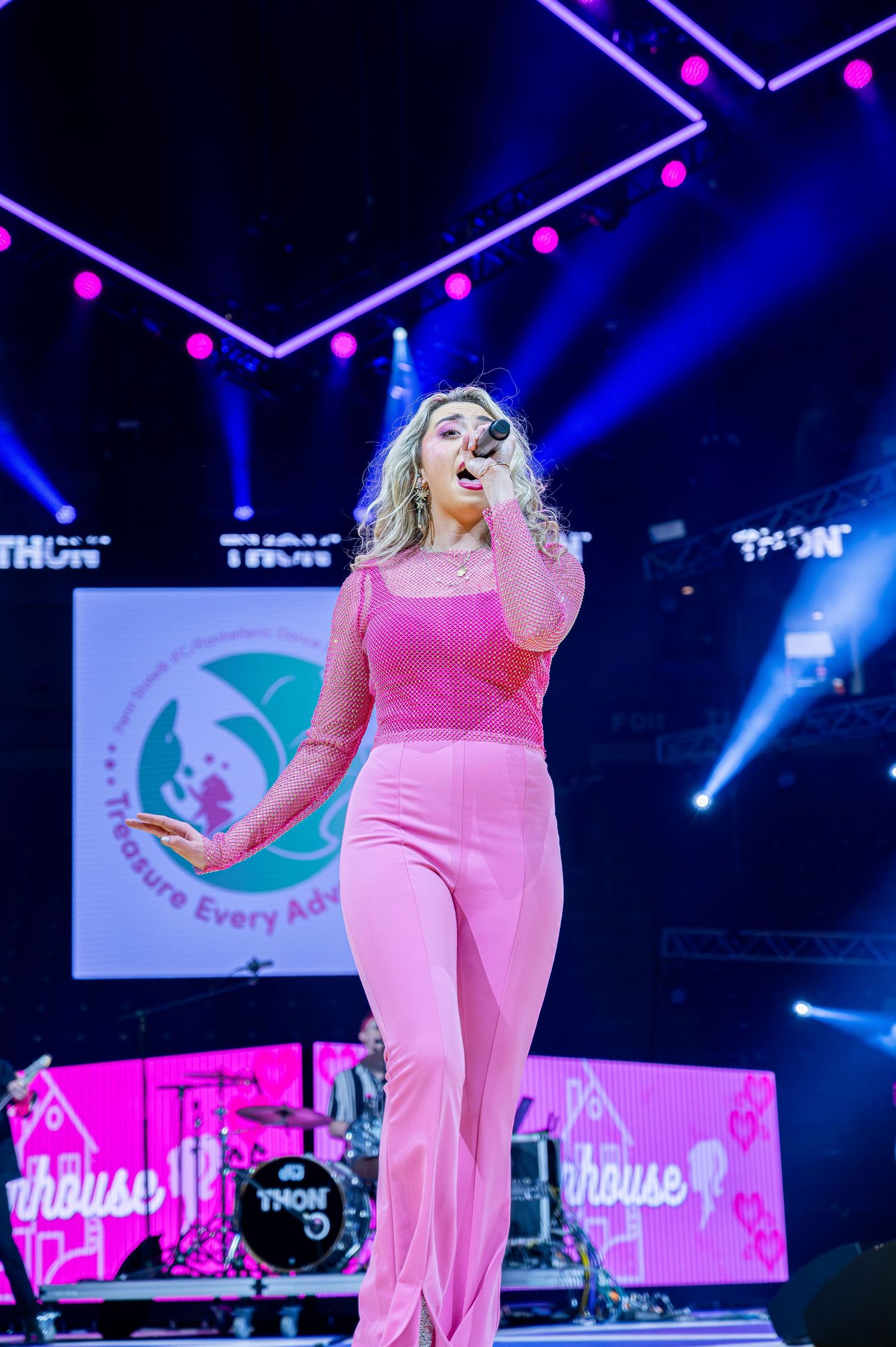 A performer in a bright pink outfit sings on stage with dramatic blue lighting and geometric patterns in the background.