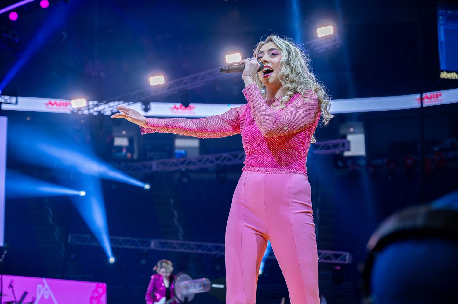 A performer in a bright pink outfit sings passionately on stage with blue lighting and spotlights in the background.