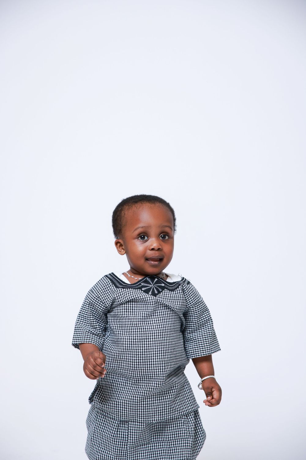 A toddler in a checkered grey outfit smiles at the camera against a white background.