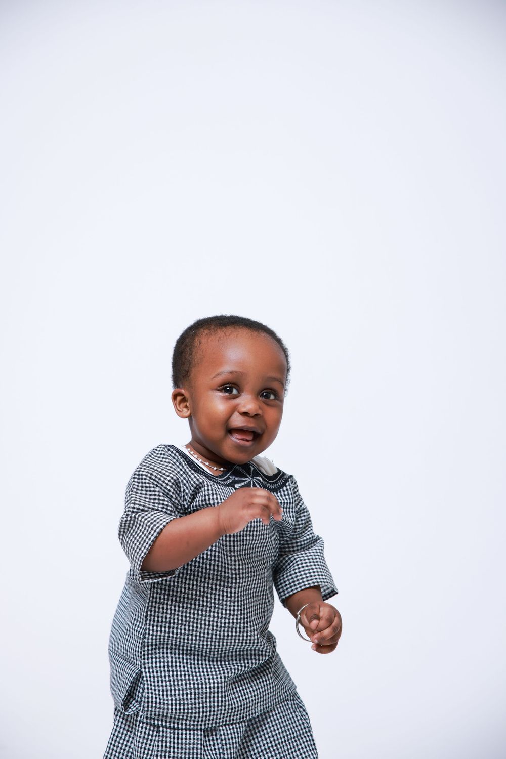 A toddler in a grey checkered outfit points while smiling against a white backdrop.