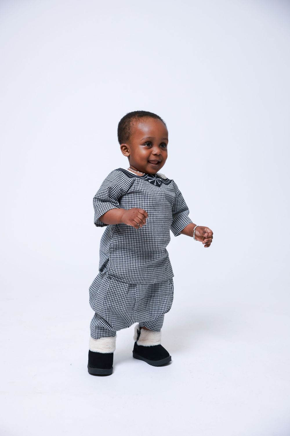 A happy toddler in a grey checkered outfit takes steps against a white background.