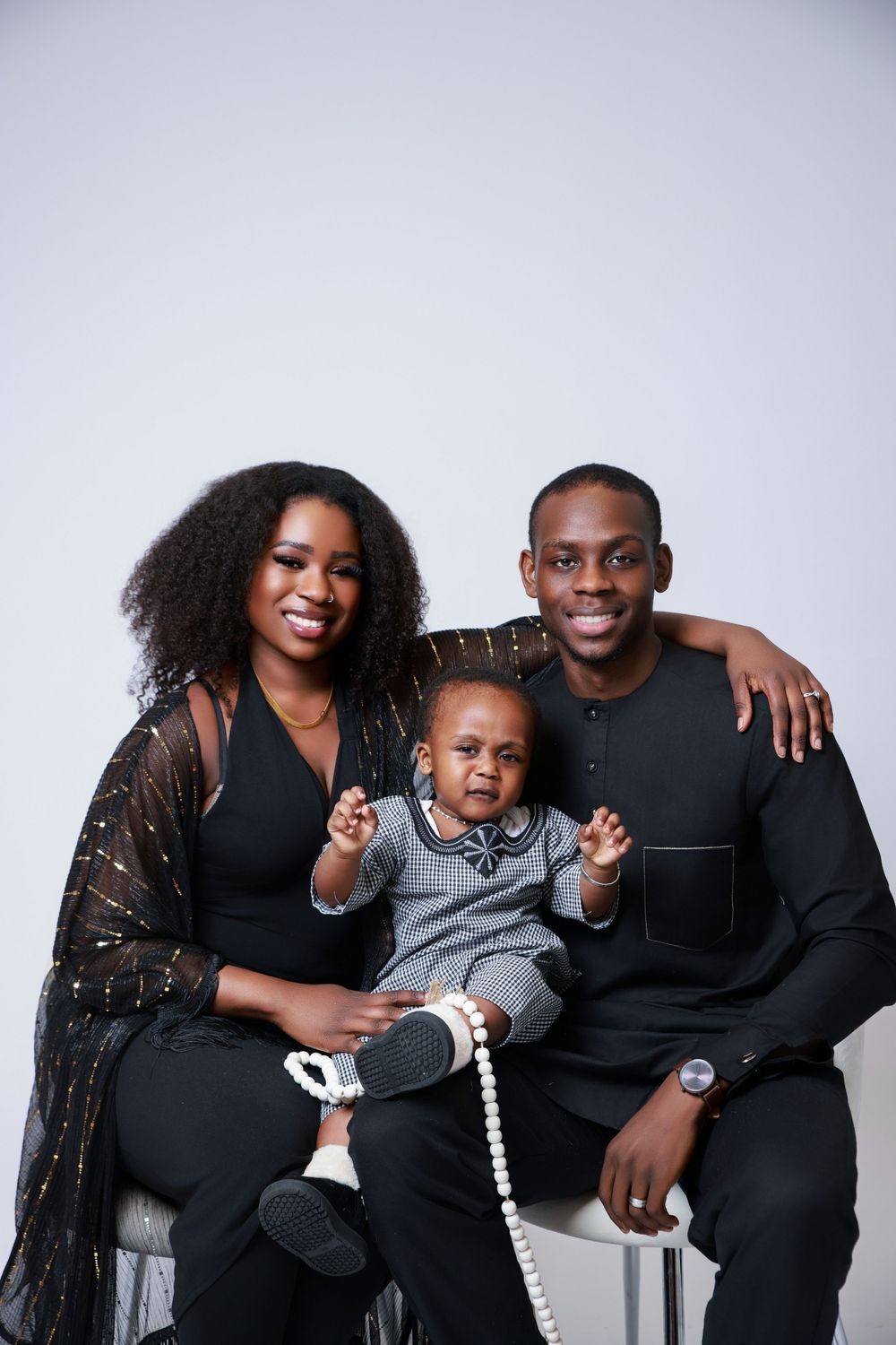 A joyful family portrait with parents and toddler sitting together against a light background.