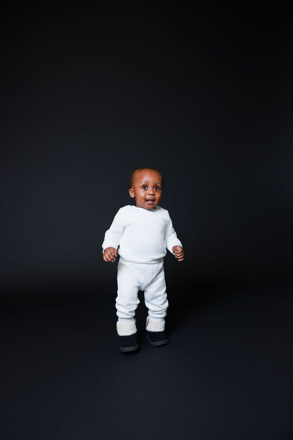 Toddler in all-white outfit standing against dark background.