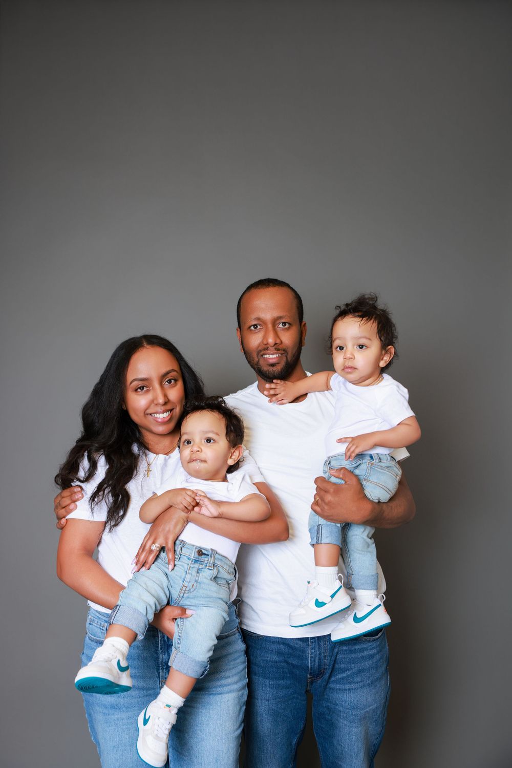 Family of four wearing coordinating white tops and jeans pose together against grey background.
