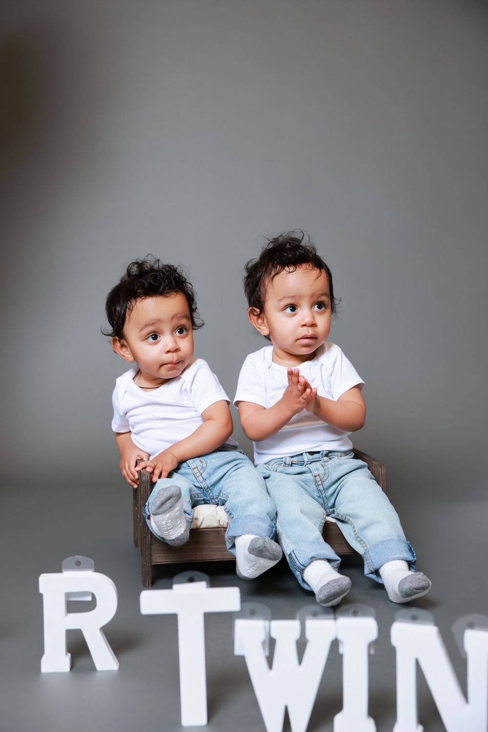 Twin toddlers in white shirts and jeans sit together with letter decorations spelling 'TWIN'.