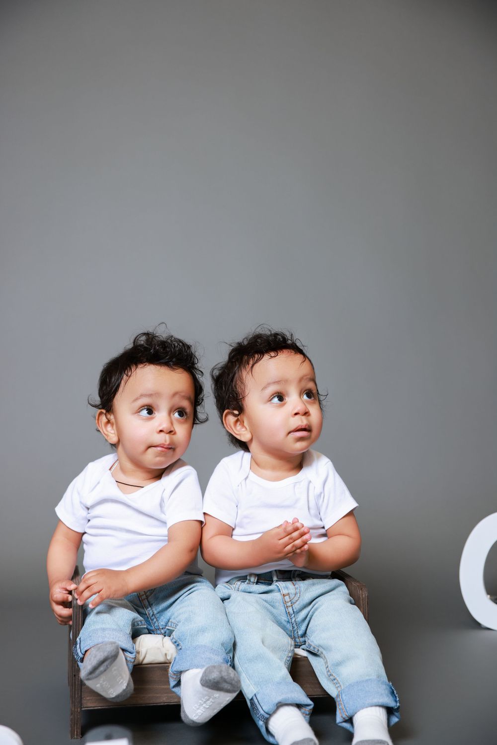 Two toddlers in matching white shirts and jeans sit side by side looking off camera.