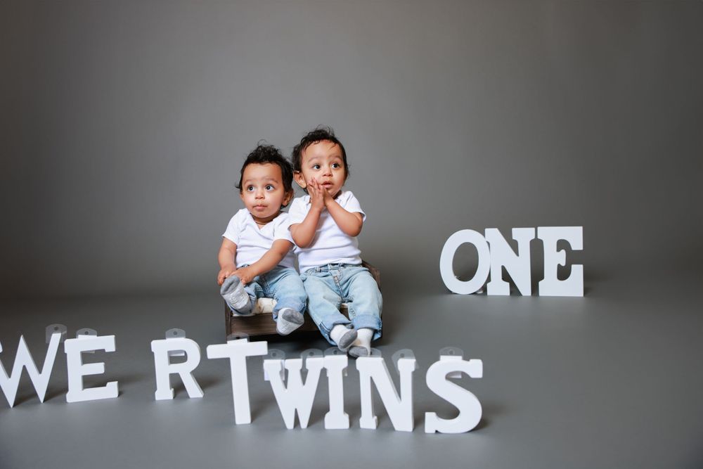 Twin babies sit together in front of a grey wall with text reading "WE RT TWINS ONE".