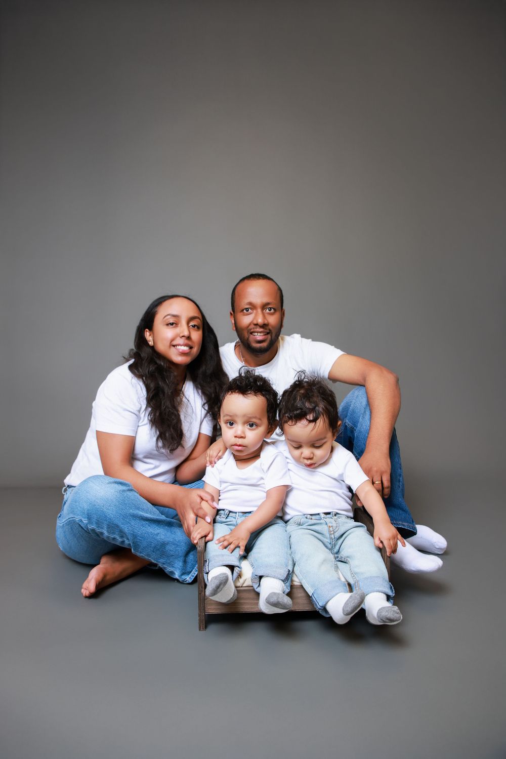Family with twins poses together on floor against grey backdrop in coordinated casual outfits.