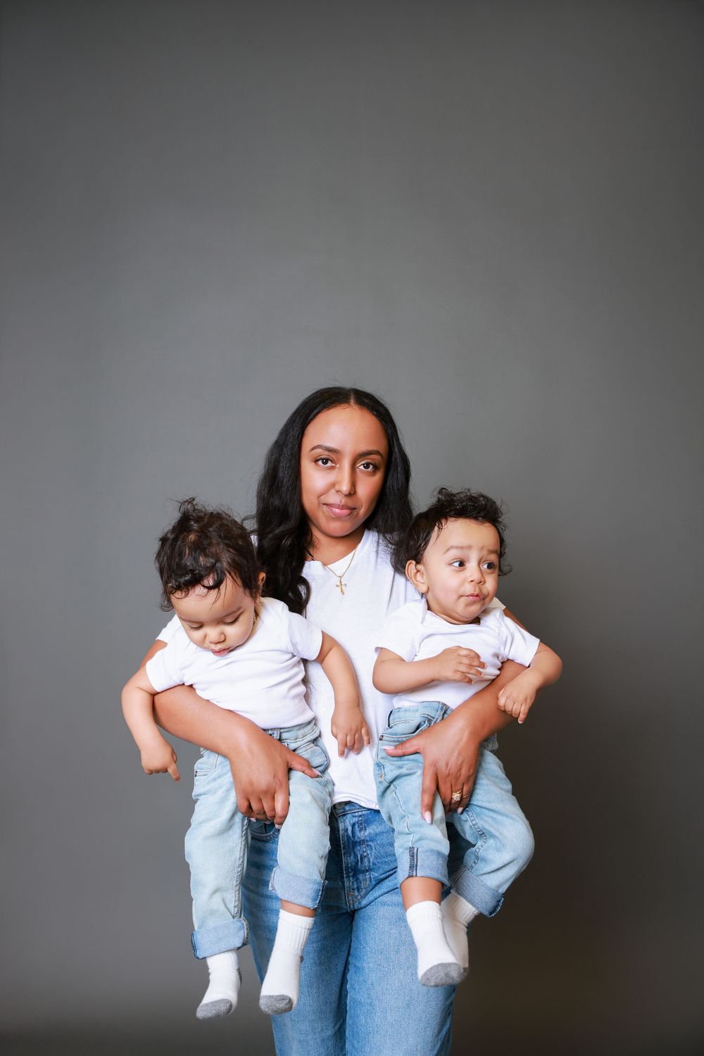Parent holding two toddlers in matching outfits poses against grey background.