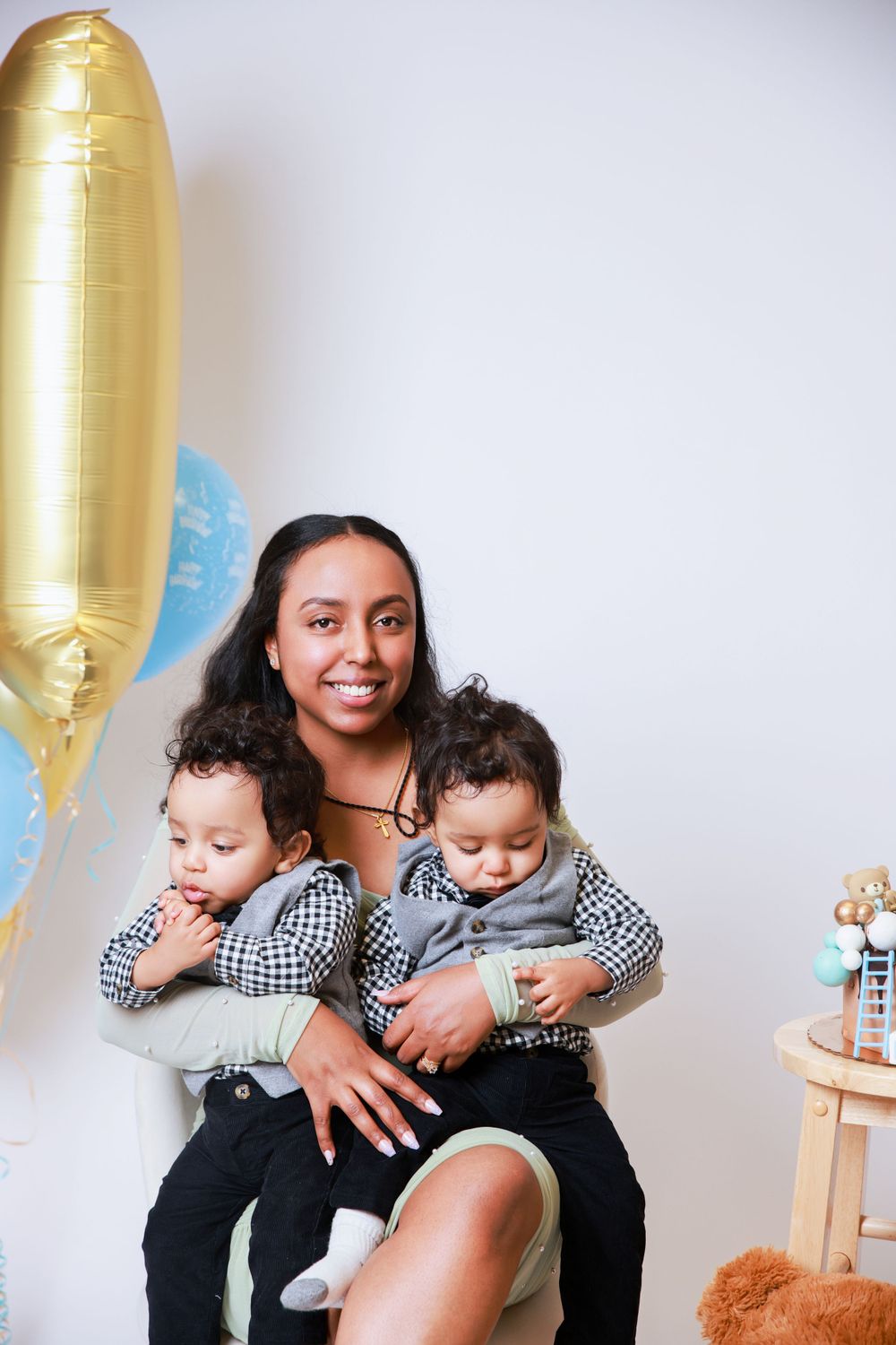 Parent holds two toddlers in matching checkered shirts with festive gold balloon decoration.