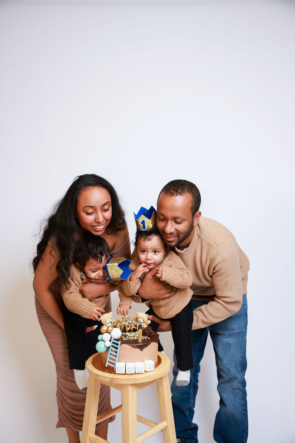 A family leans in together around a decorated birthday cake on a wooden stool.