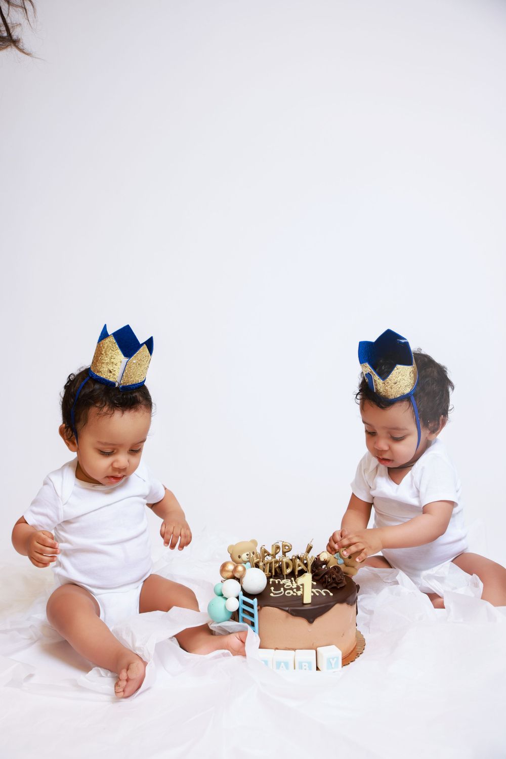 Two babies in white outfits and gold crowns examine a birthday cake together.