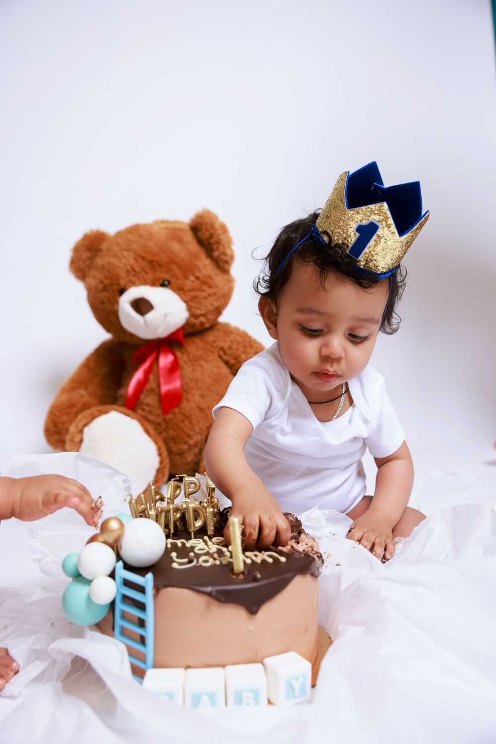 A baby in a crown touches a birthday cake while a teddy bear sits nearby.