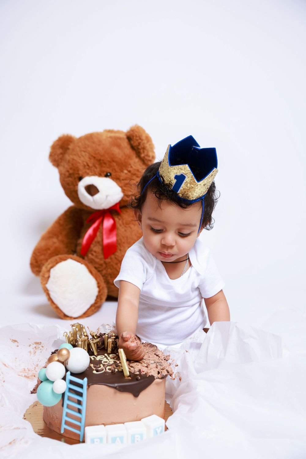 A baby in white wearing a blue and gold crown reaches for a chocolate birthday cake.