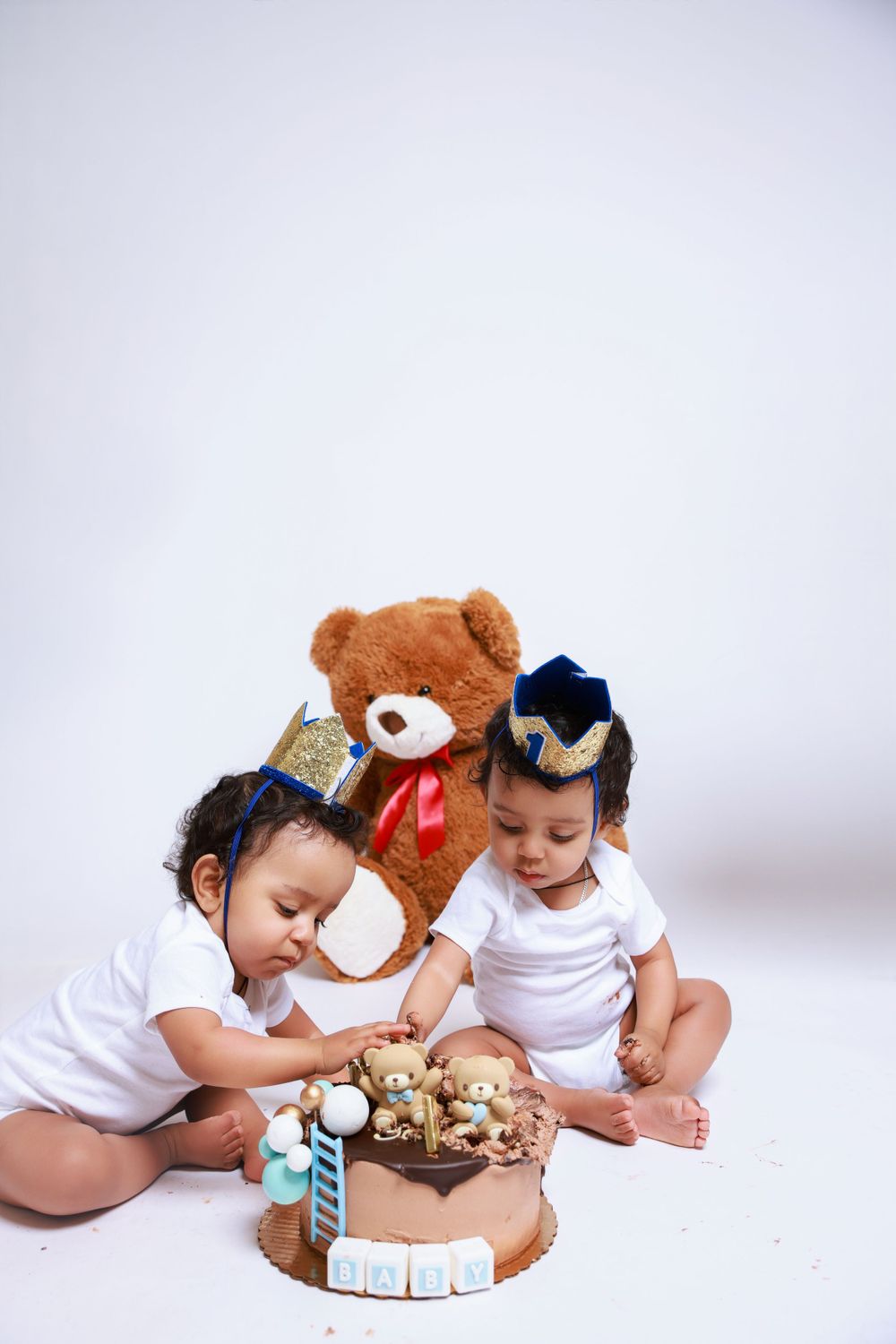 Two babies explore a birthday cake together next to a brown teddy bear.