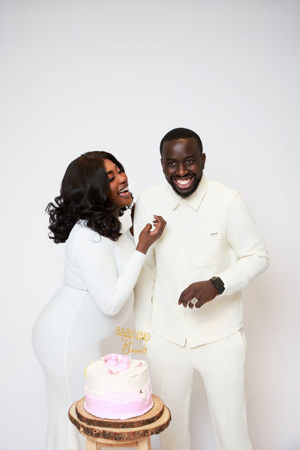 Couple in white attire sharing a playful moment with a pink cake during their photoshoot.
